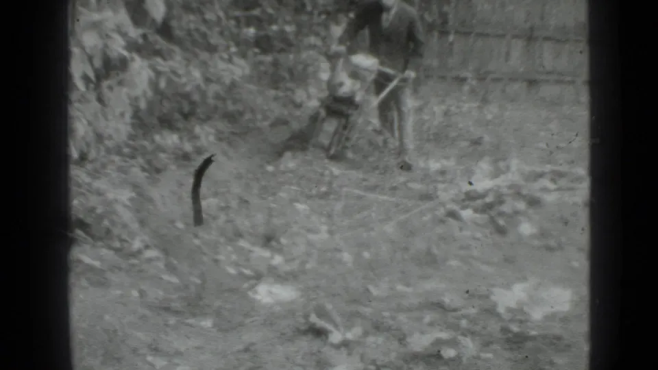 1975 Moscow-area 8mm frame showing a man guiding a small hand tractor through uneven soil near vegetation and a fence.