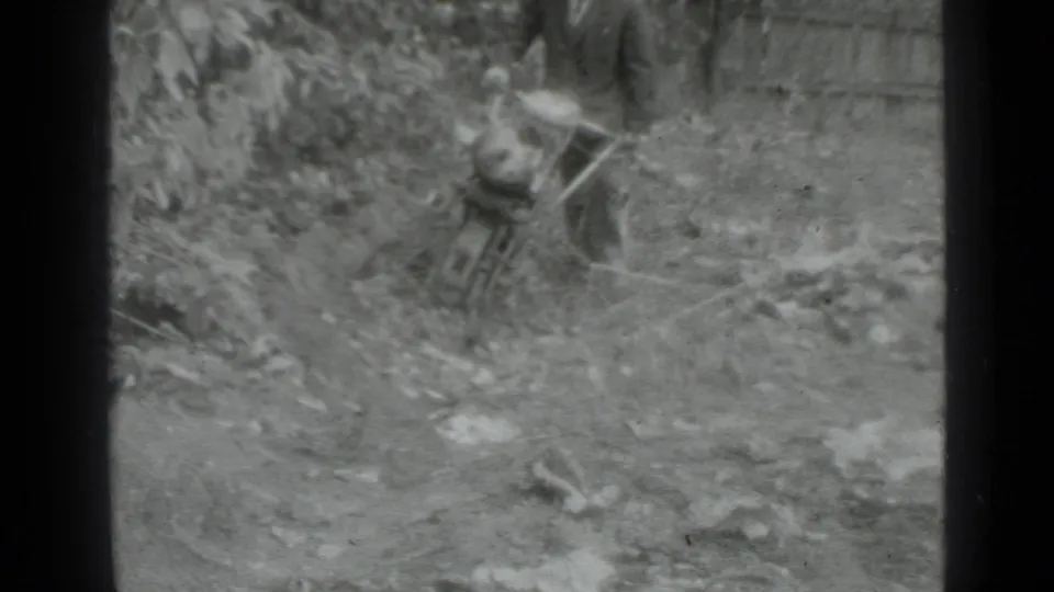 Soft black-and-white 8mm frame showing the man with the hand tractor in the same rough garden setting near vegetation.