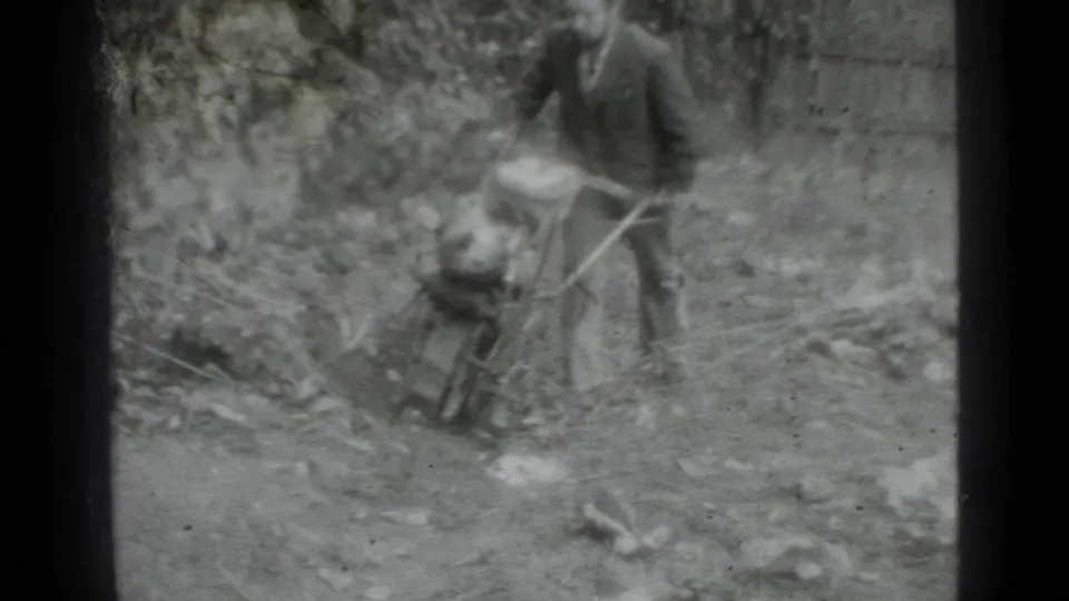 Closer 1975 Moscow home-movie frame with the man leaning into a small hand tractor on uneven garden ground.