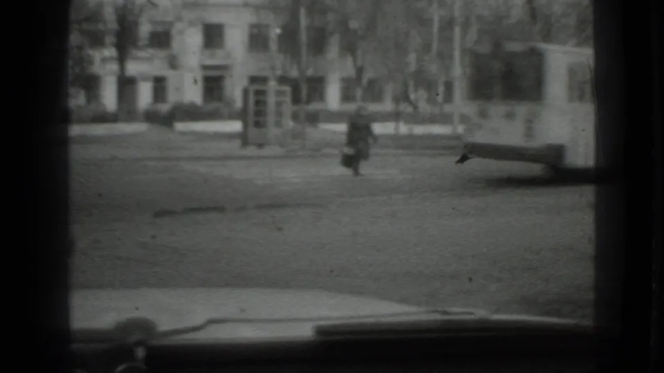 Moscow 1975 home-movie frame showing a distant woman crossing an open street, viewed through dark vehicle-window framing.