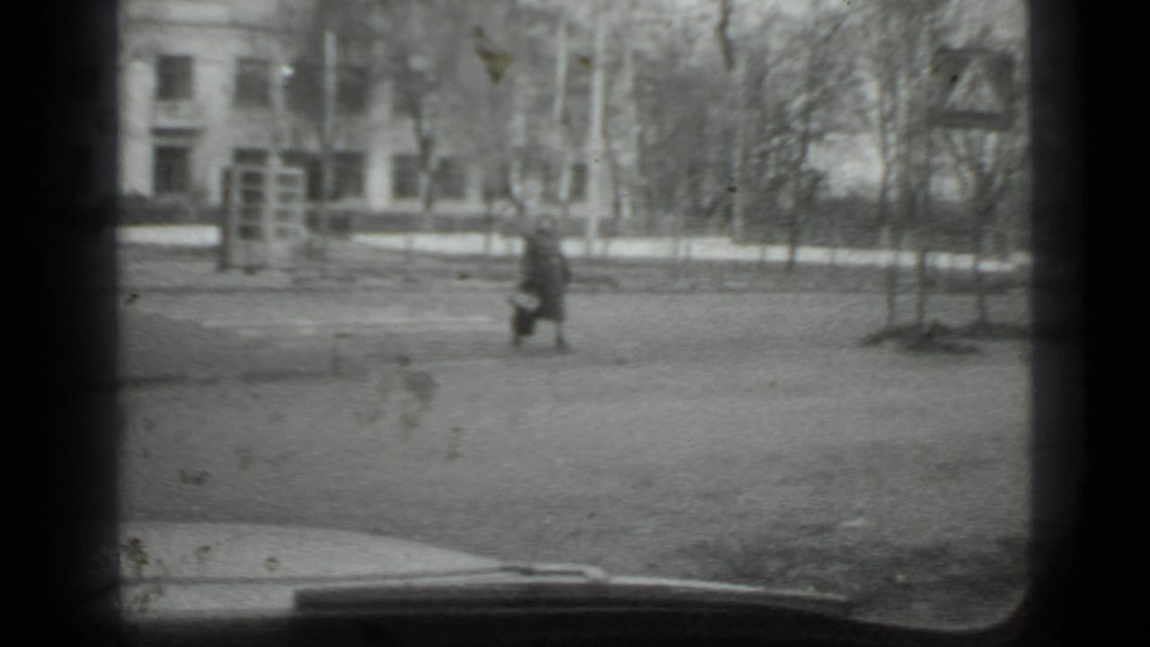 Woman walking near a roadside sign with buildings and leafless trees behind her.