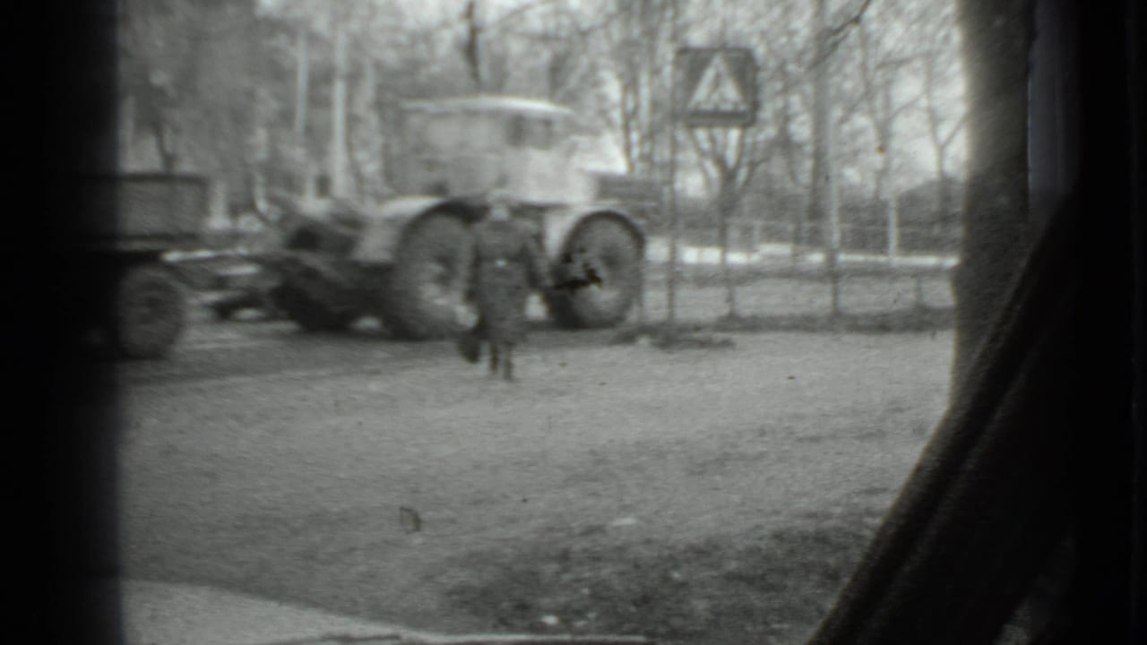1975 Moscow home-movie frame showing a woman in a long coat near a truck and trailer, with bare trees and window edges visible.