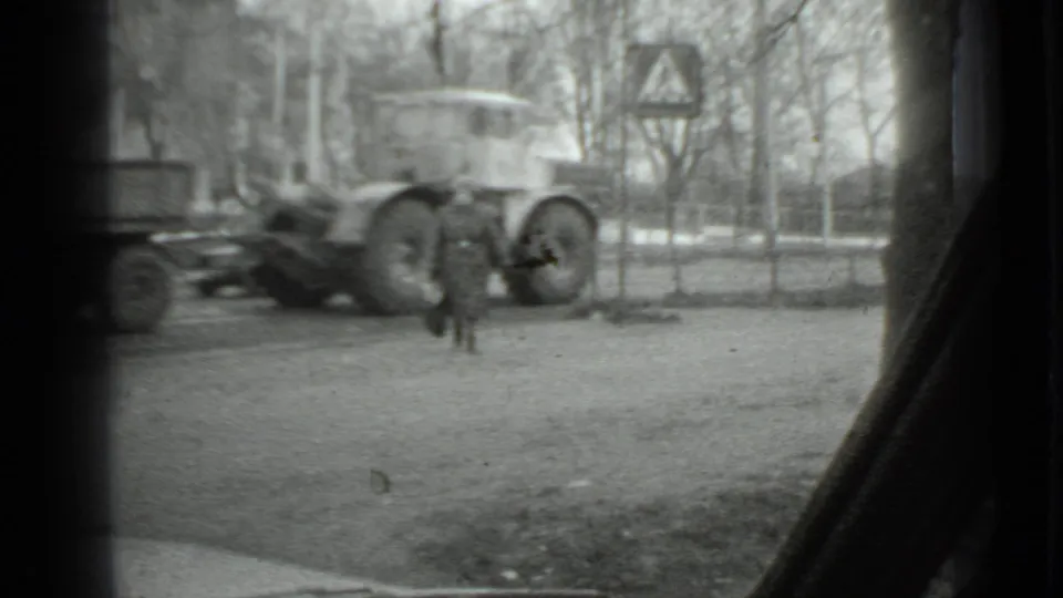 1975 Moscow home-movie frame showing a woman in a long coat near a truck and trailer, with bare trees and window edges visible.