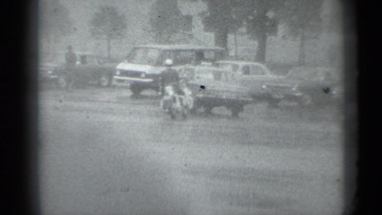 Black-and-white 1983 Moscow street view with pedestrians, a motorcycle, and vintage cars moving or parked near trees and buildings.