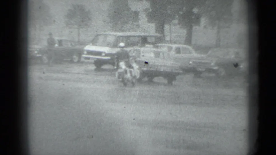 Black-and-white 1983 Moscow street view with pedestrians, a motorcycle, and vintage cars moving or parked near trees and buildings.