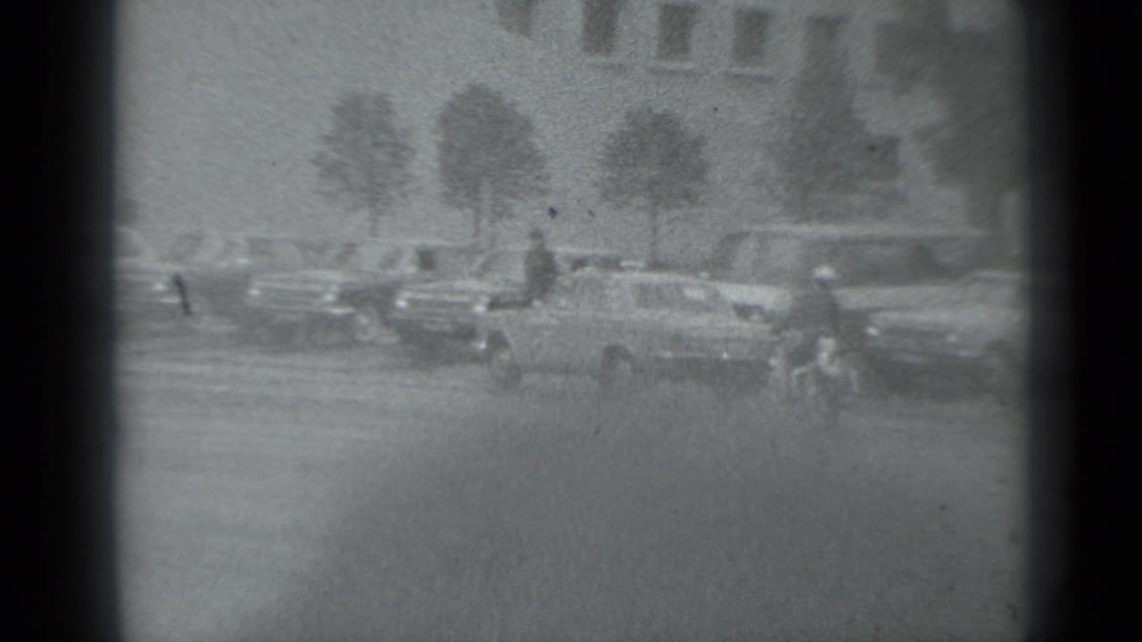 Wide street view with multiple cars, a pedestrian, trees, and a large building in the background.