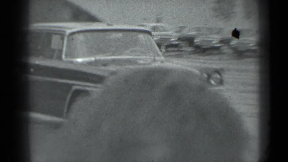 Soft black-and-white 8mm frame with a large car rear in the foreground and blurred street traffic behind it in Moscow.