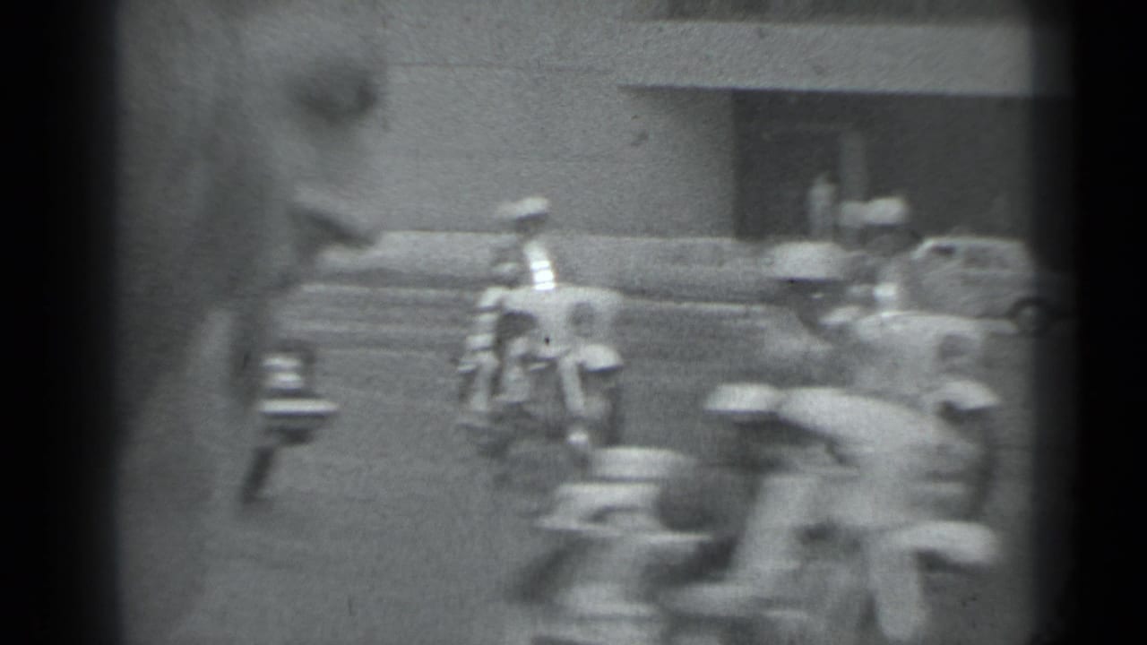 Motorcycles and cars pass through a street scene, partly blurred by movement.