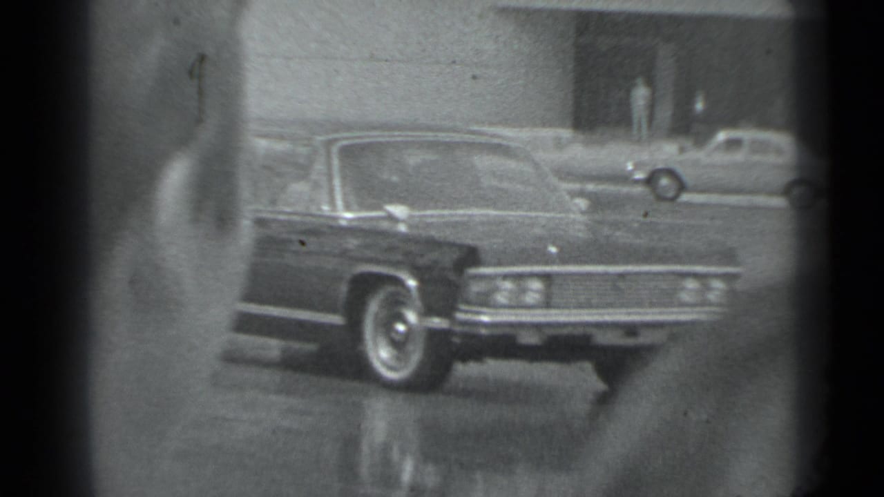 Dark sedan passing on a city street, partly obscured by film gate edges.
