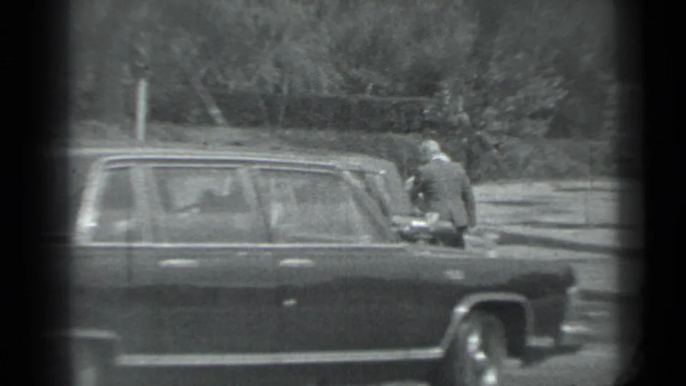 A dark sedan is seen side-on near a curb in Moscow, with suited adults standing behind it in grainy 1983 8mm footage.