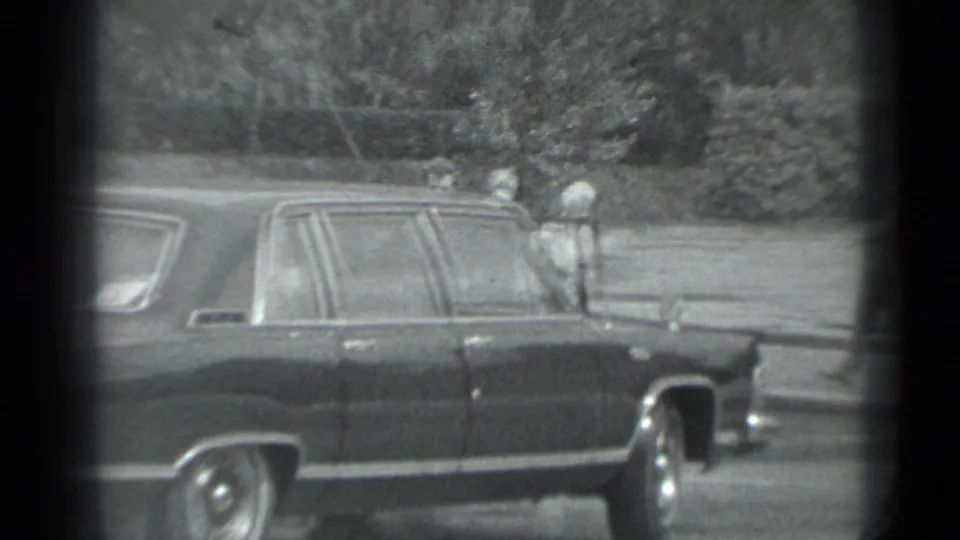 Side view of the dark sedan in 1983 Moscow, with several adults visible around the far side of the car and a leafy background.