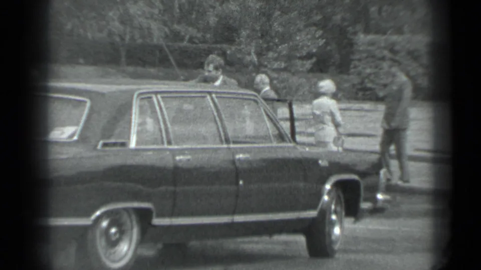 1983 Moscow home-movie frame showing adults around a dark sedan, including a light-coated woman and suited men near the curb.
