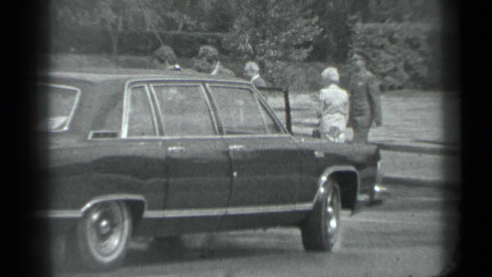 1983 Moscow home-movie frame showing a dark sedan near bystanders in a public parking or curbside area.