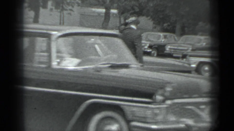Moscow 1983 frame showing the front of a dark car crossing the view, with older vehicles parked along the street.