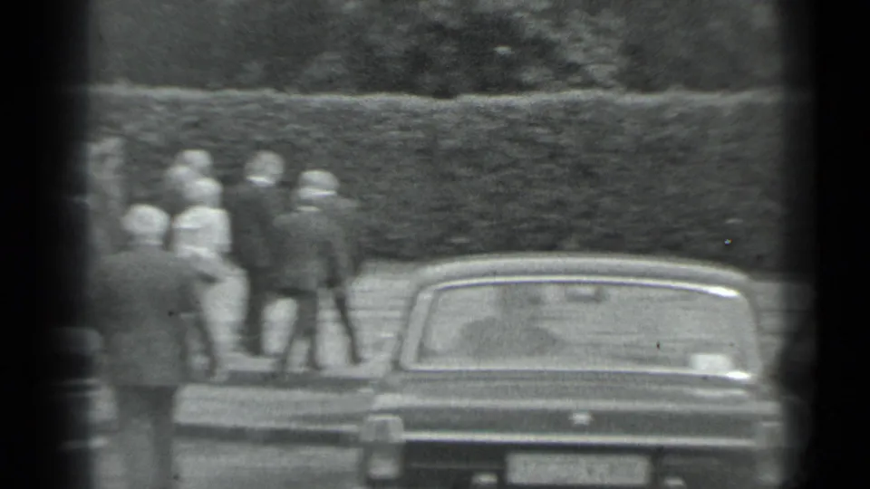 Black-and-white 8mm frame of suited men and a woman walking away near a parked car and hedge in Moscow, 1983.