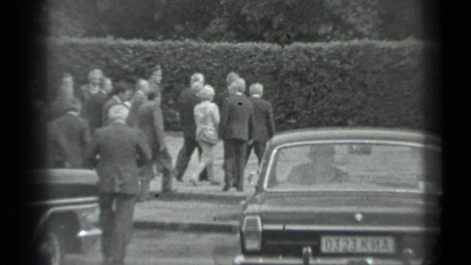 A larger formal group gathers beside a hedge and vintage car, with one woman visible among suited men in the 1983 Moscow footage.
