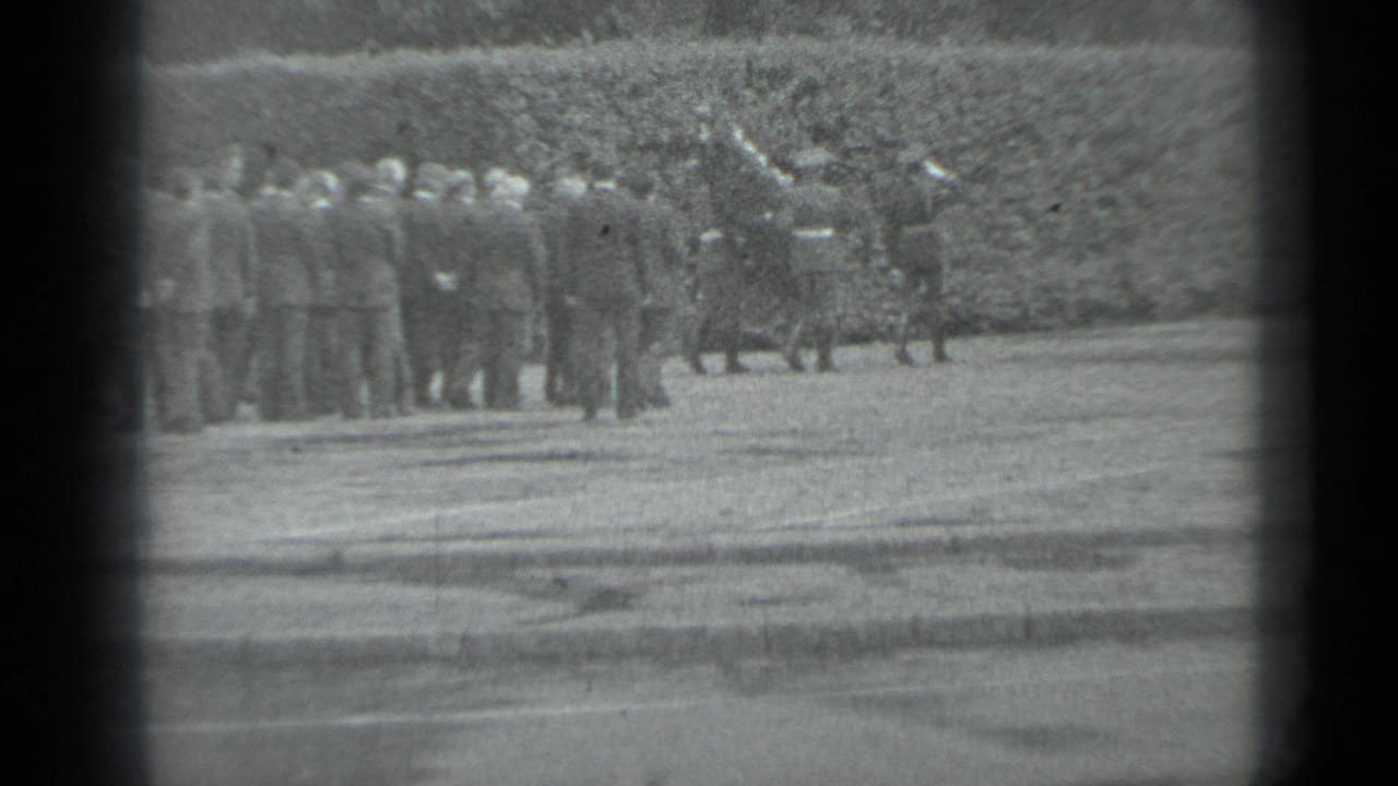 Distant adults in formal clothing gather beside a hedge during the supplied 1983 Moscow funeral procession footage.