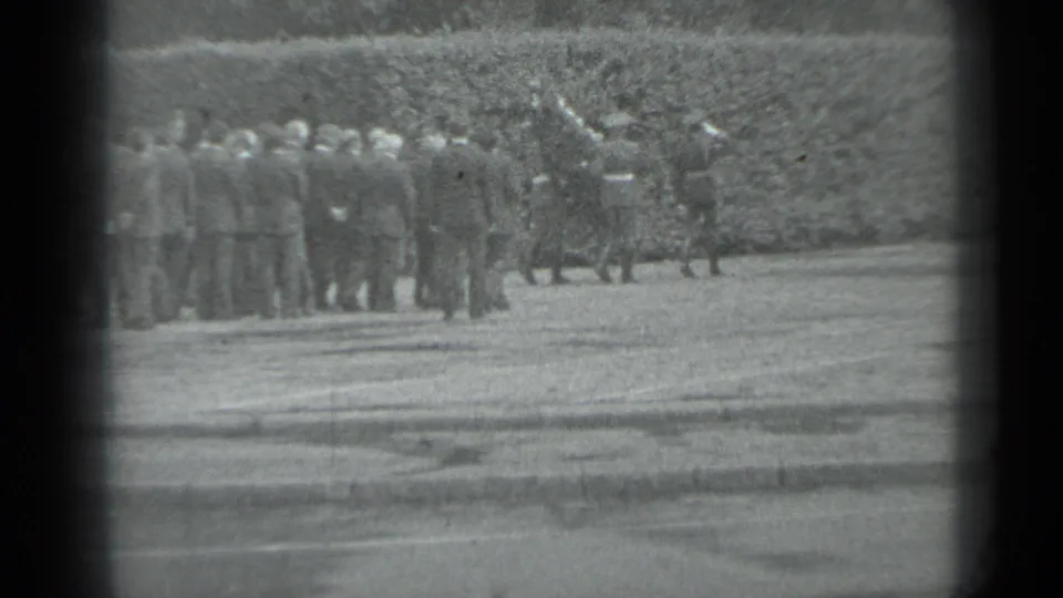Distant adults in formal clothing gather beside a hedge during the supplied 1983 Moscow funeral procession footage.