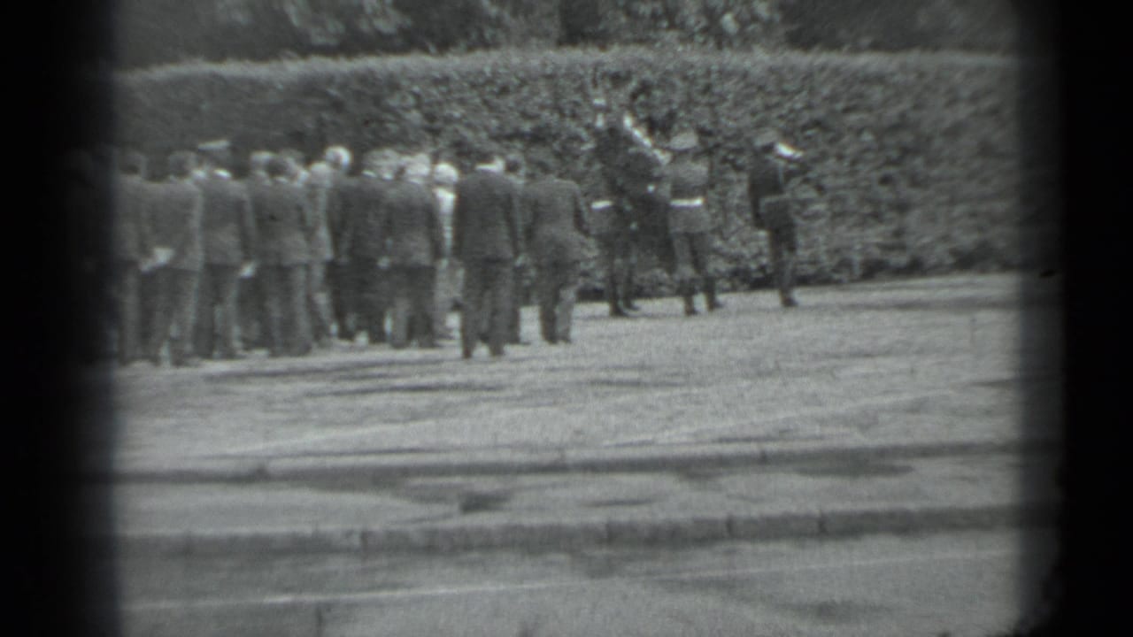 Adults in formal attire stand near a hedge in the supplied Moscow funeral procession clip, filmed from a distance.