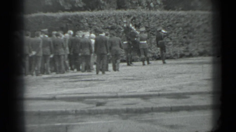 Adults in formal attire stand near a hedge in the supplied Moscow funeral procession clip, filmed from a distance.