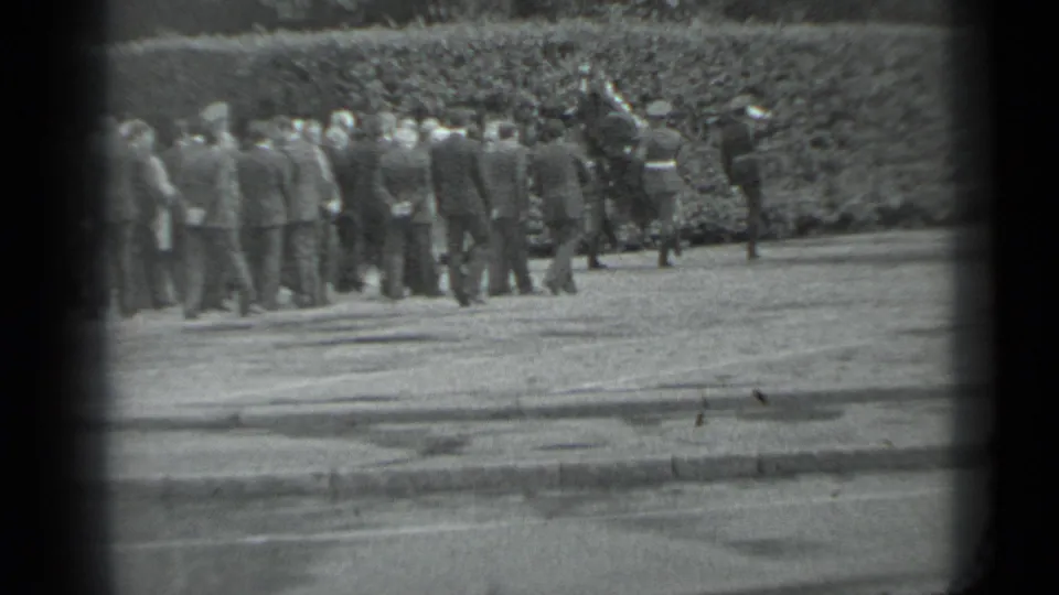 Mourners walk in formation beside a hedge in black-and-white 8mm footage identified as Moscow in 1983.