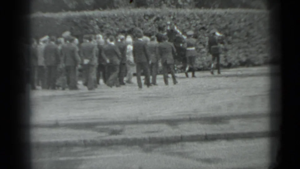 Black-and-white 8mm frame of adults in suits gathered outdoors in Moscow, with a hedge and a few distant uniformed figures visible.