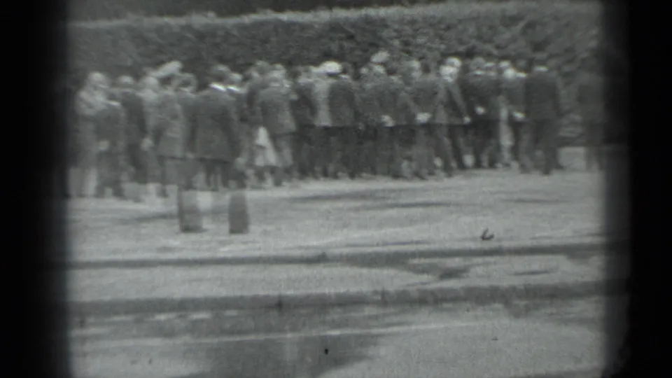 Black-and-white Moscow frame of a formal outdoor crowd near a hedge, with foreground pavement and short posts visible.