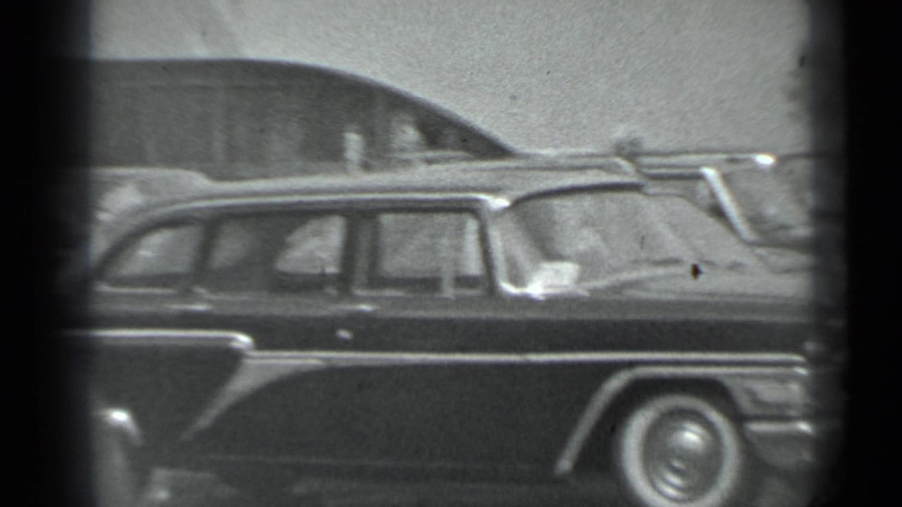 Black-and-white 8mm frame of a large dark mid-century sedan seen from the side at the Moscow car display.