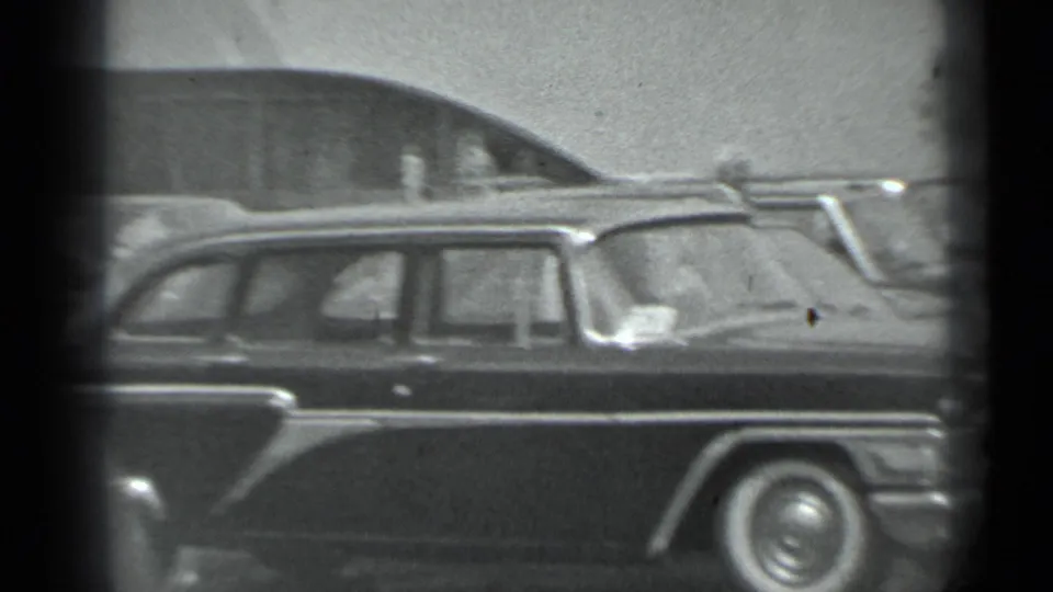 Black-and-white 8mm frame of a large dark mid-century sedan seen from the side at the Moscow car display.