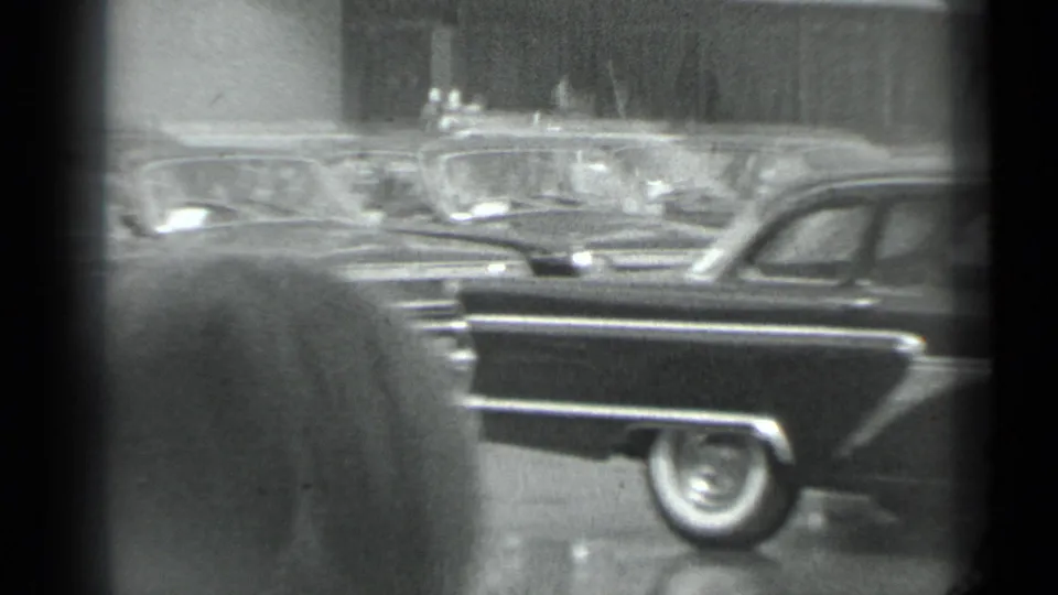 Visitor-shot 1983 Moscow frame with a spectator partly blocking the view of a dark classic sedan and parked cars behind it.