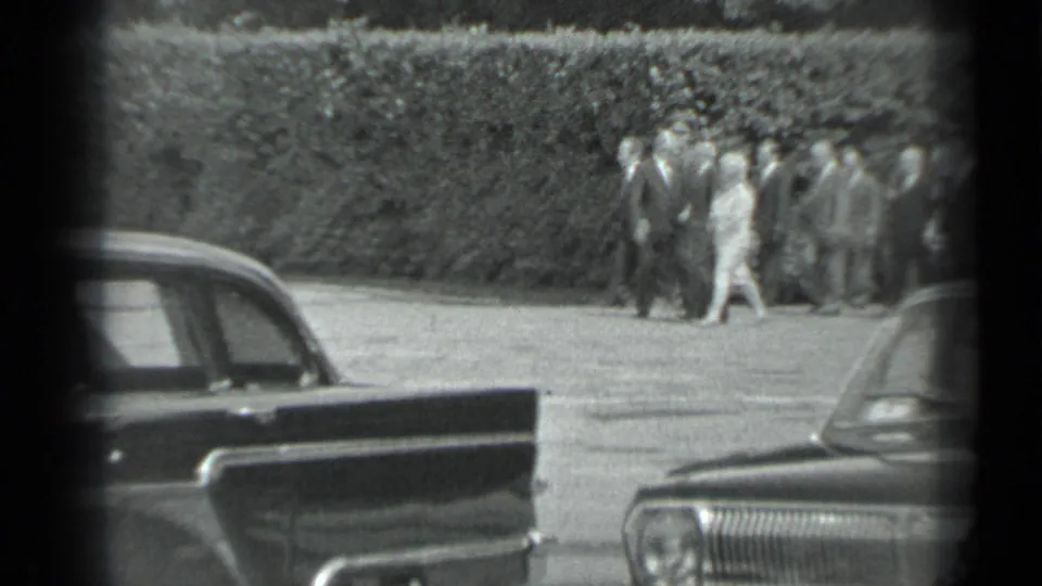 1983 Moscow 8mm frame showing formally dressed pedestrians near a hedge, including one woman, with dark parked cars in front.