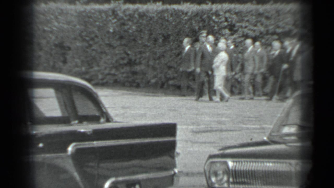 In the supplied 1983 Moscow context, a formally dressed group walks across paved grounds near parked sedans and a tall hedge.
