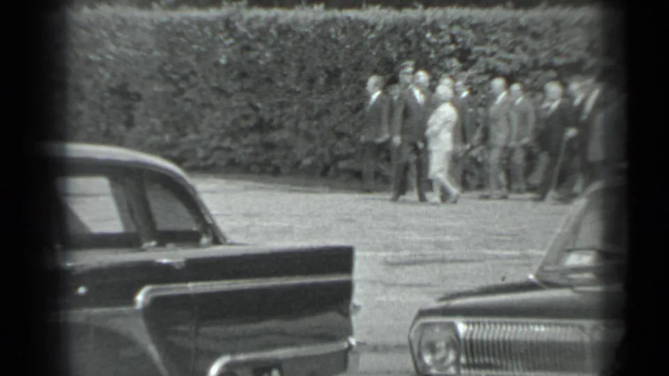 In the supplied 1983 Moscow context, a formally dressed group walks across paved grounds near parked sedans and a tall hedge.