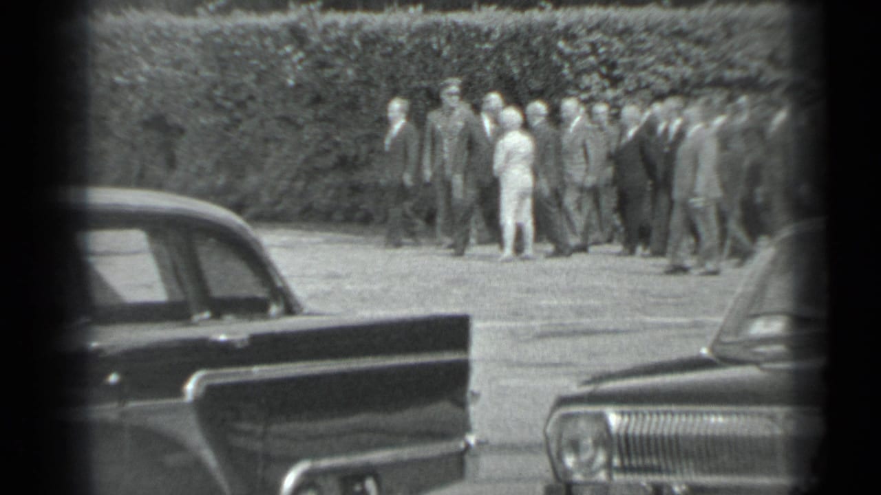 A group of adults walks and talks outdoors, partly framed by parked cars.