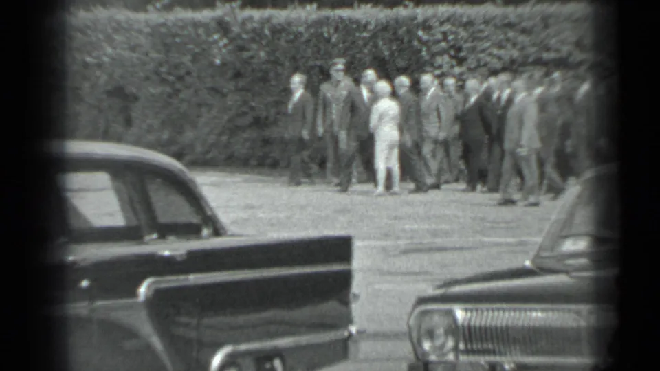 The group continues walking in the supplied Moscow setting, with a light-dressed woman among suited men and sedans in the foreground.