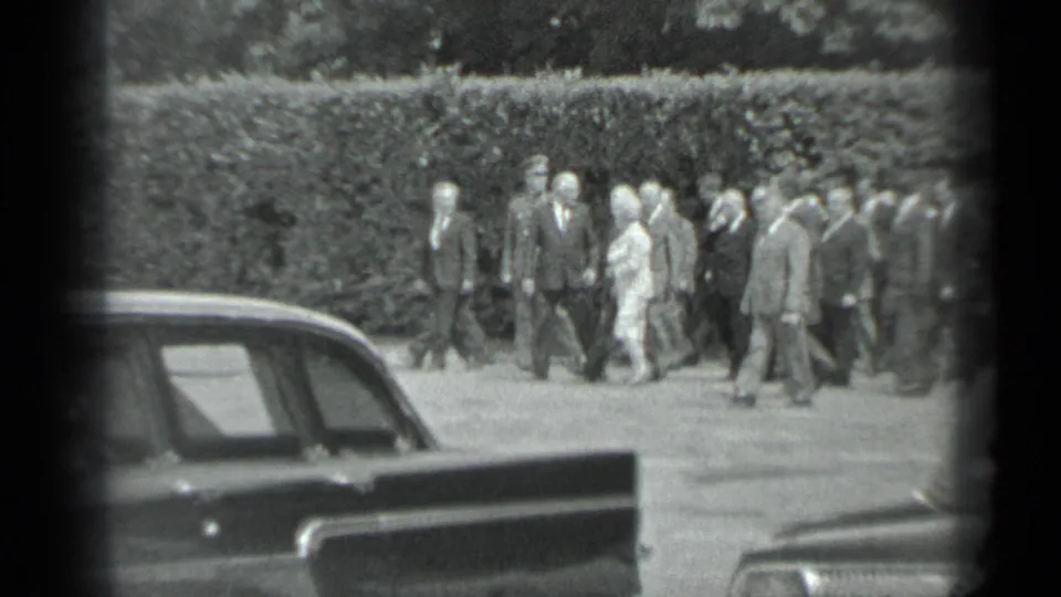 A formal group walks across the frame in the 1983 Moscow footage, with parked cars close to the camera and a hedge behind them.