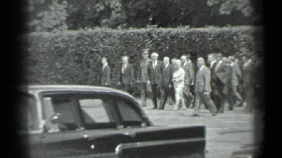 The group appears closer together near the end of the Moscow clip, walking past a parked sedan with hedge-lined grounds behind them.