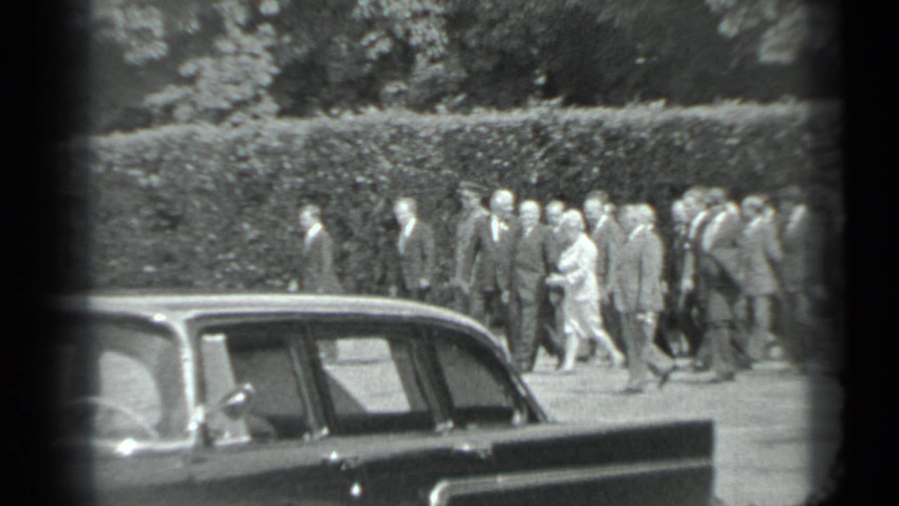 In 1983 Moscow 8mm footage, a formal group walks along a park path behind a dark parked sedan and hedge.