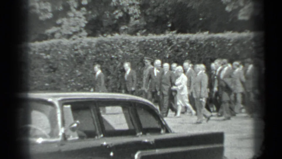 In 1983 Moscow 8mm footage, a formal group walks along a park path behind a dark parked sedan and hedge.