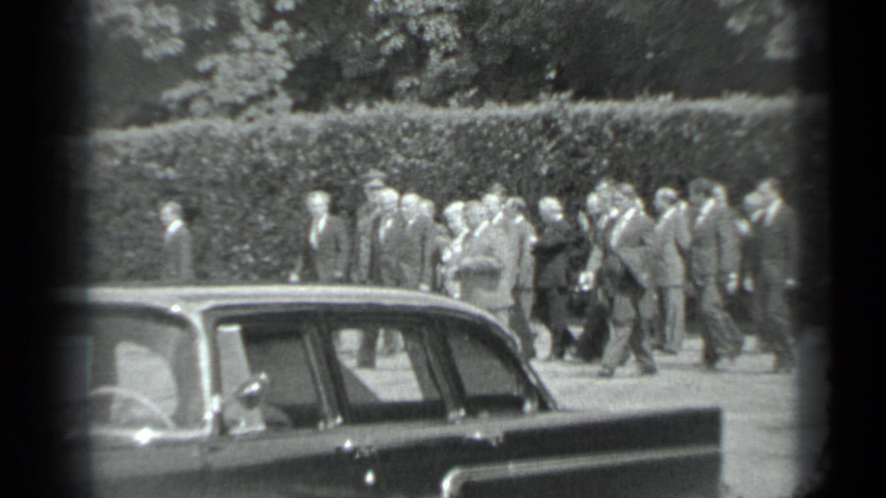 The Moscow park procession continues, with suited men and women in dresses moving in formation behind a parked sedan.