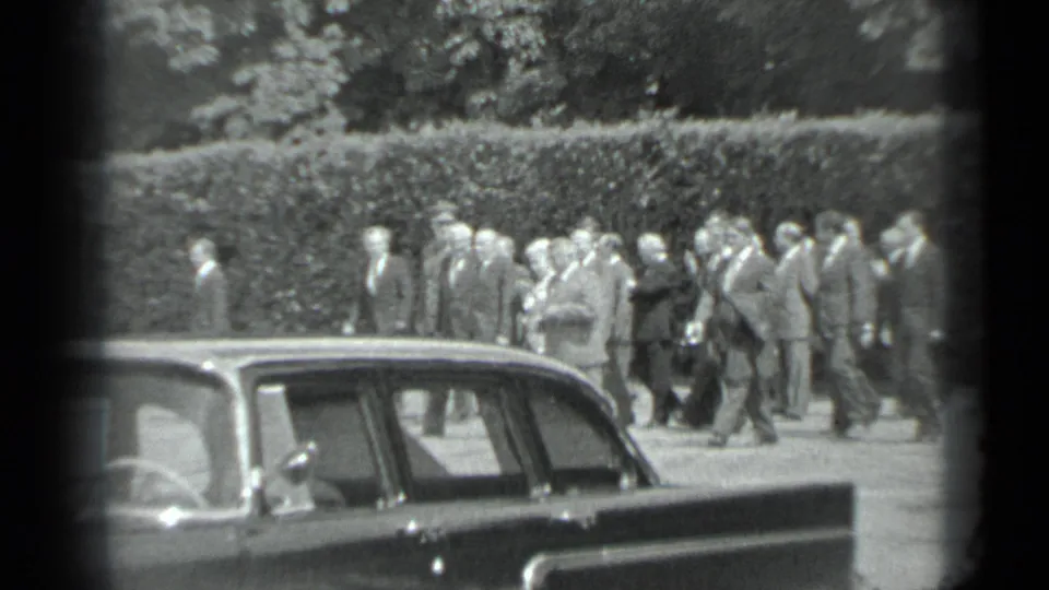 The Moscow park procession continues, with suited men and women in dresses moving in formation behind a parked sedan.