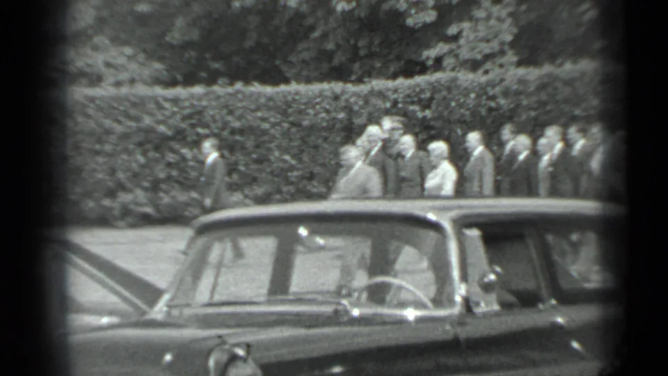 Near the end of the 1983 Moscow clip, people in formal clothing appear beyond a parked sedan with a hedge and trees behind them.