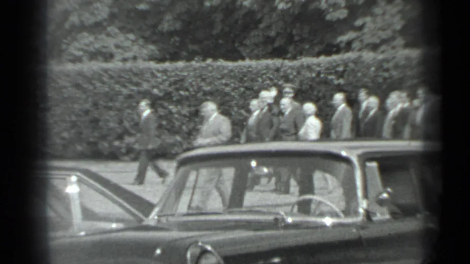 In 1983 Moscow footage, a formally dressed group walks past a hedge while car roofs and windows partly block the view.