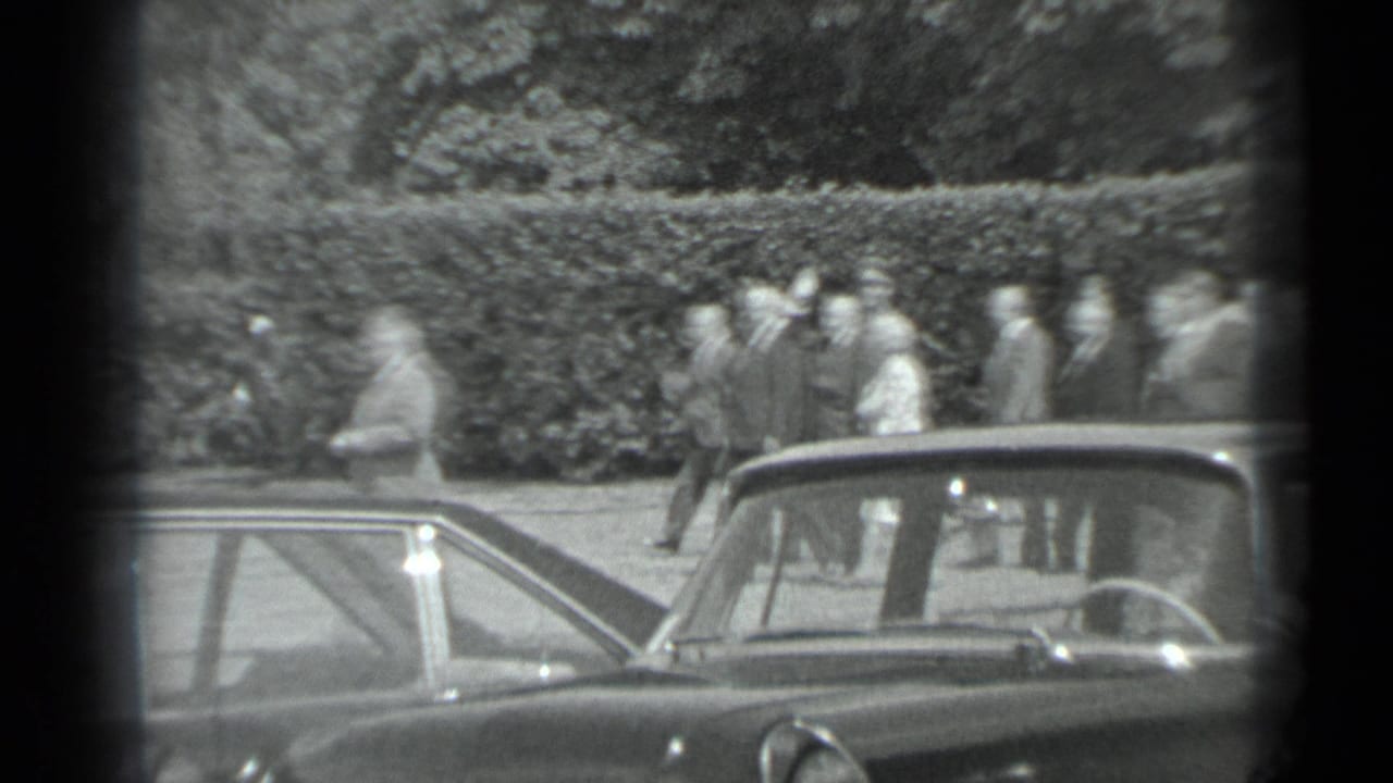 A distant group in suits and light clothing moves along a hedge in Moscow, partly obscured by a dark frame edge and car rooflines.