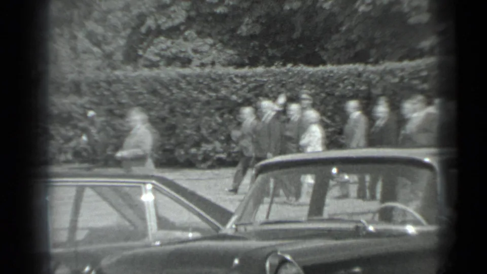 A distant group in suits and light clothing moves along a hedge in Moscow, partly obscured by a dark frame edge and car rooflines.