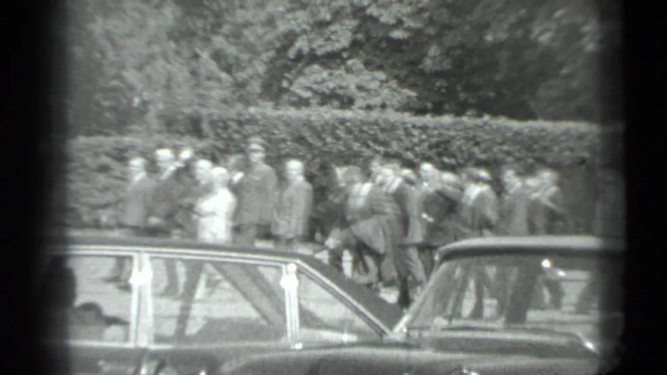 Black-and-white 1983 Moscow home-movie frame showing suited adults and a light-dressed woman near parked cars and hedges.