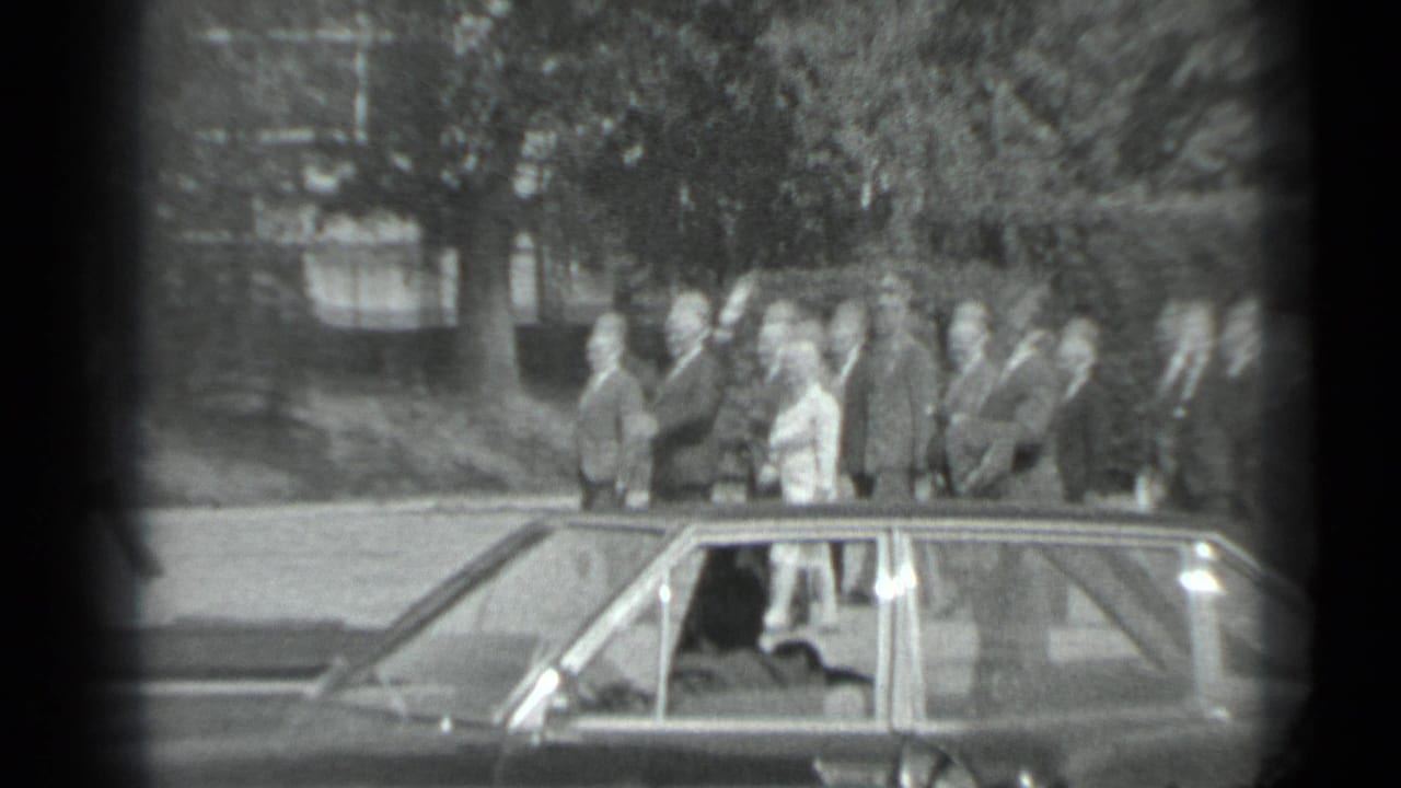 Black-and-white Moscow frame with formally dressed adults near a sedan, including a man appearing to wave or gesture.