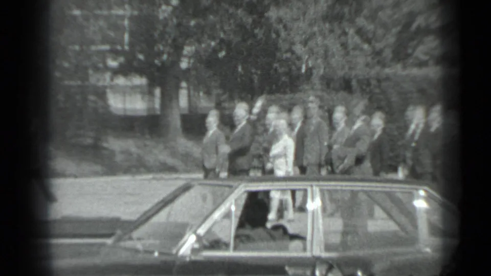 Black-and-white Moscow frame with formally dressed adults near a sedan, including a man appearing to wave or gesture.