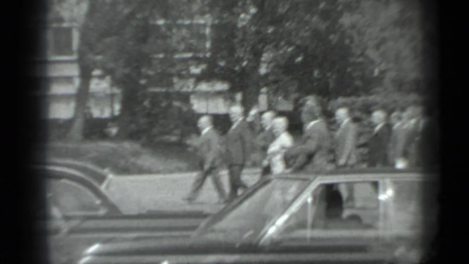 Black-and-white 1983 Moscow frame showing adults walking together past parked cars in a tree-lined area.