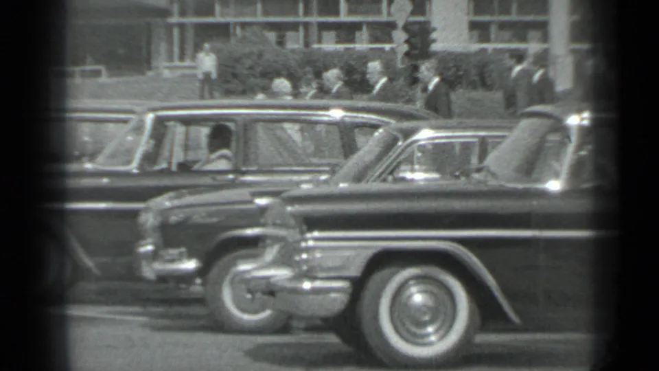 Black-and-white Moscow home-movie frame with parked period cars in front and adults gathered near a building in the distance.